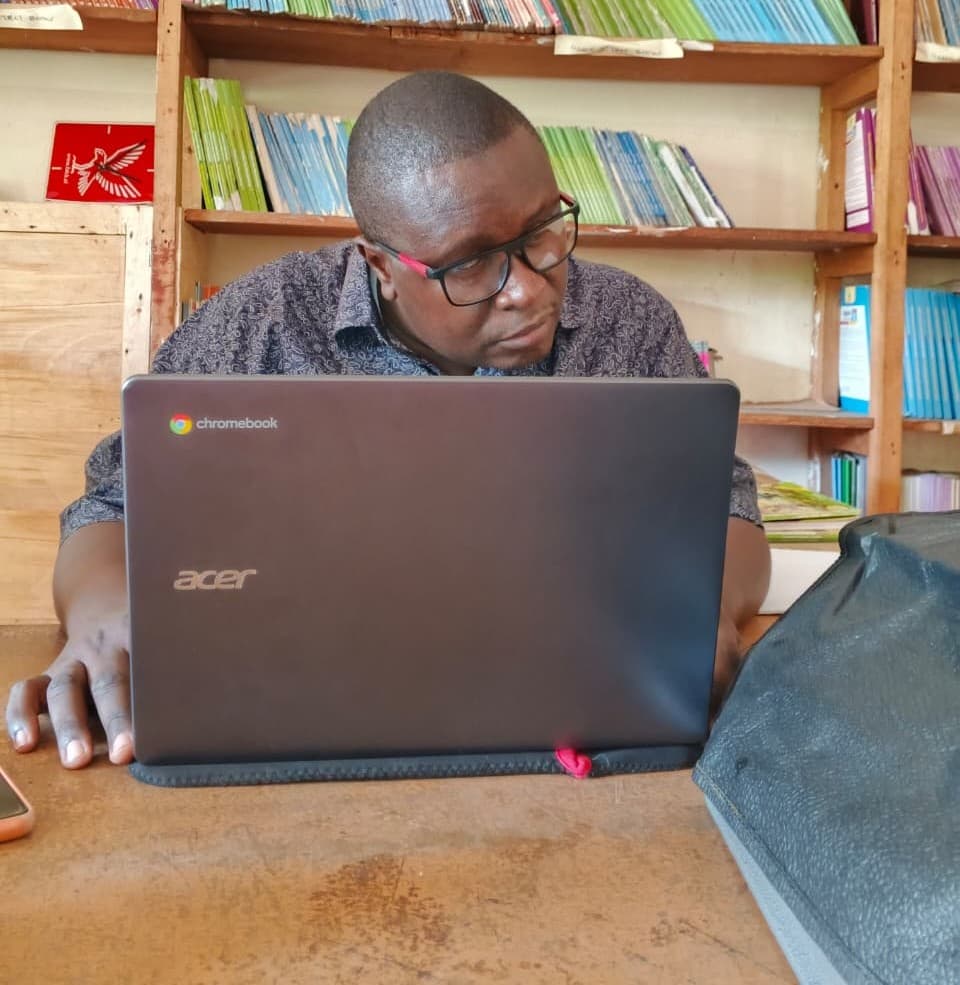 Mr Sammy works on an Acer Chromebook at a desk in a library or resource room with shelves of colorful books in the background.