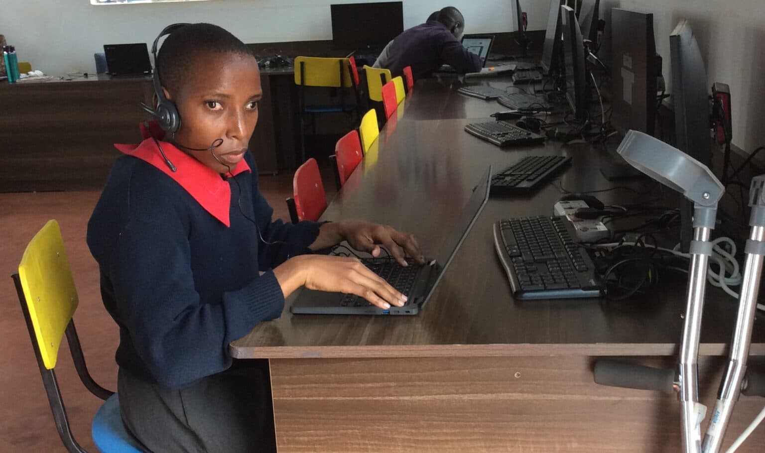 Janet sits at a computer workstation in an inABLE computer lab, using a laptop with a headset and microphone. Several desktop computers are arranged along a long table, while another students works at a computer in the background. A pair of forearm crutch