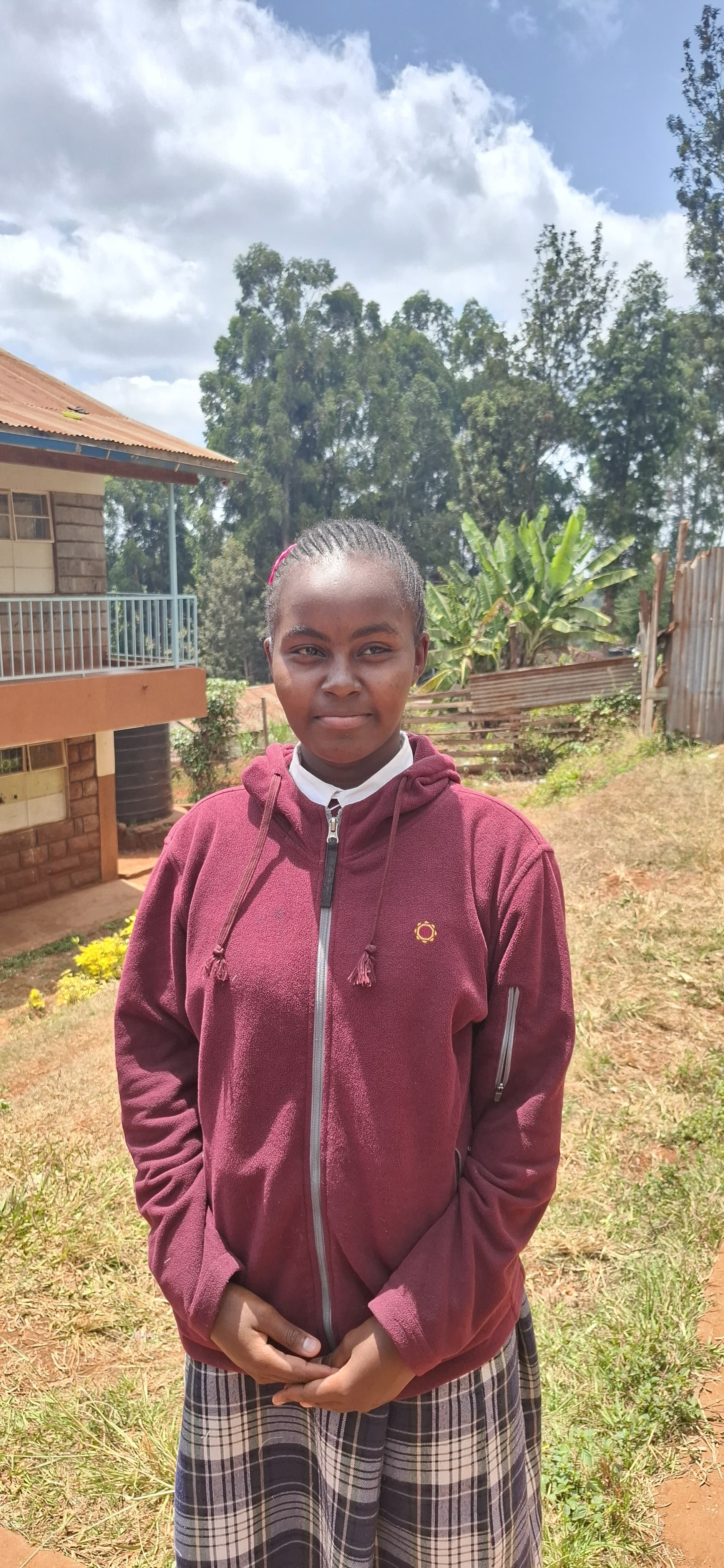 Sharin stands outdoors on a grassy school compound, wearing a maroon zip-up school sweater over a white collared shirt and a plaid skirt. The student is facing the camera with hands clasped in front.