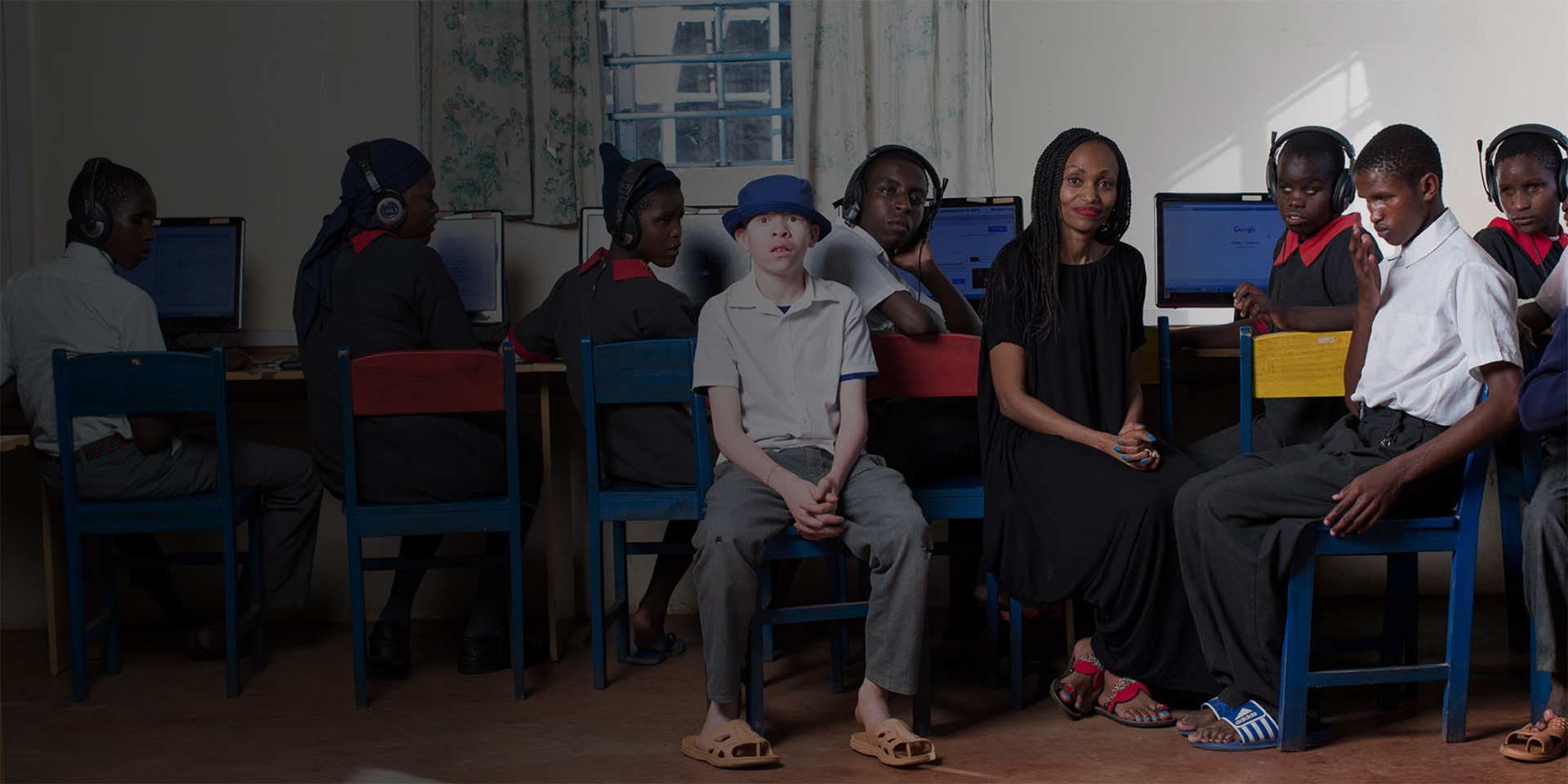 Irene Mbari-Kirika, Founder & Executive Director of inABLE smiles at the camera while seated next to students in an accessibility lab at Thika Primary School For the Blind, Kenya.