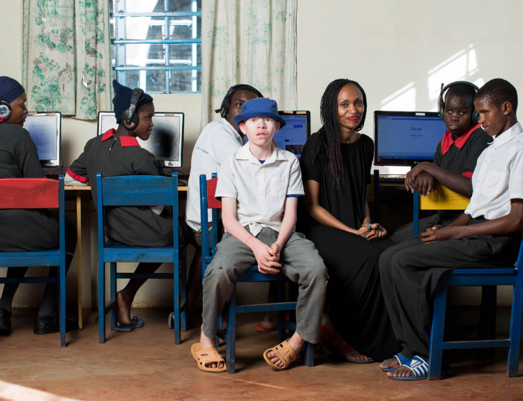 Irene Mbari-Kirika, Founder & Executive Director of inABLE smiles at the camera while seated next to students in an accessibility lab at Thika Primary School For the Blind, Kenya.