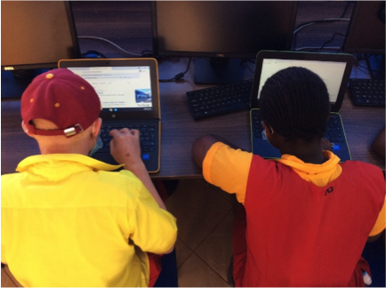 Two students use computers. The youth on the left wears a yellow shirt and red hat and types on the keyboard. On the right, the youth has dark short hair and wears red tunic with orange shirt and glances at computer screen.