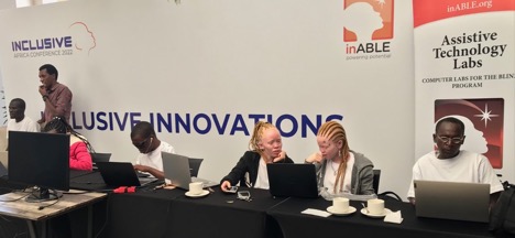 Computer coders (2 fair skinned women with braids, and 4 Black men) sit at a table with laptop computers in front of them.