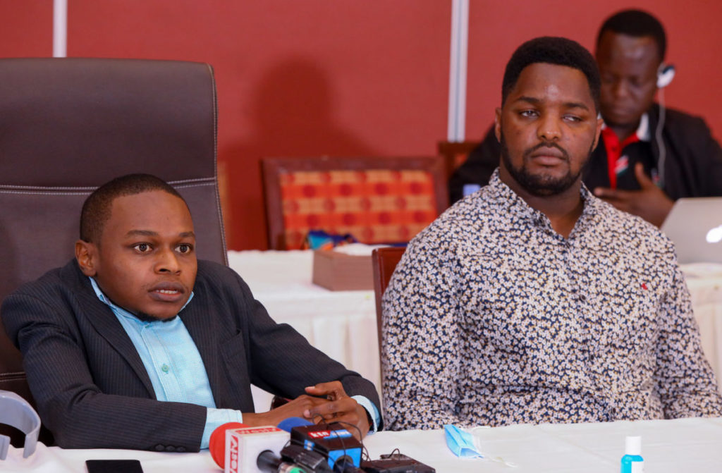 Two black men sit at a conference table looking forward. On the left sitting low is a chair is Bernard Chira wearing a blue suit. On the right is Wilson Macharia wearing a multi color dress shirt.