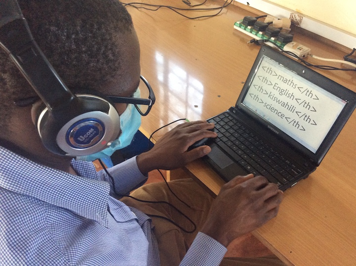Nehemiah, a black man wearing headphones, glasses, face mask and a blue dress shirt, types on a small tablet.
