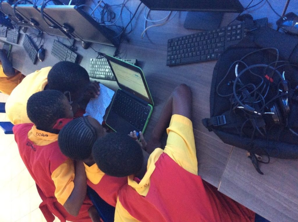 Three students dressed in yellow and red school uniform, and surrounded by keyboards and monitors, look at a laptop screen. A fourth student on the left wearing a yellow shirt closely reads from a line paper filled with handwritten words.