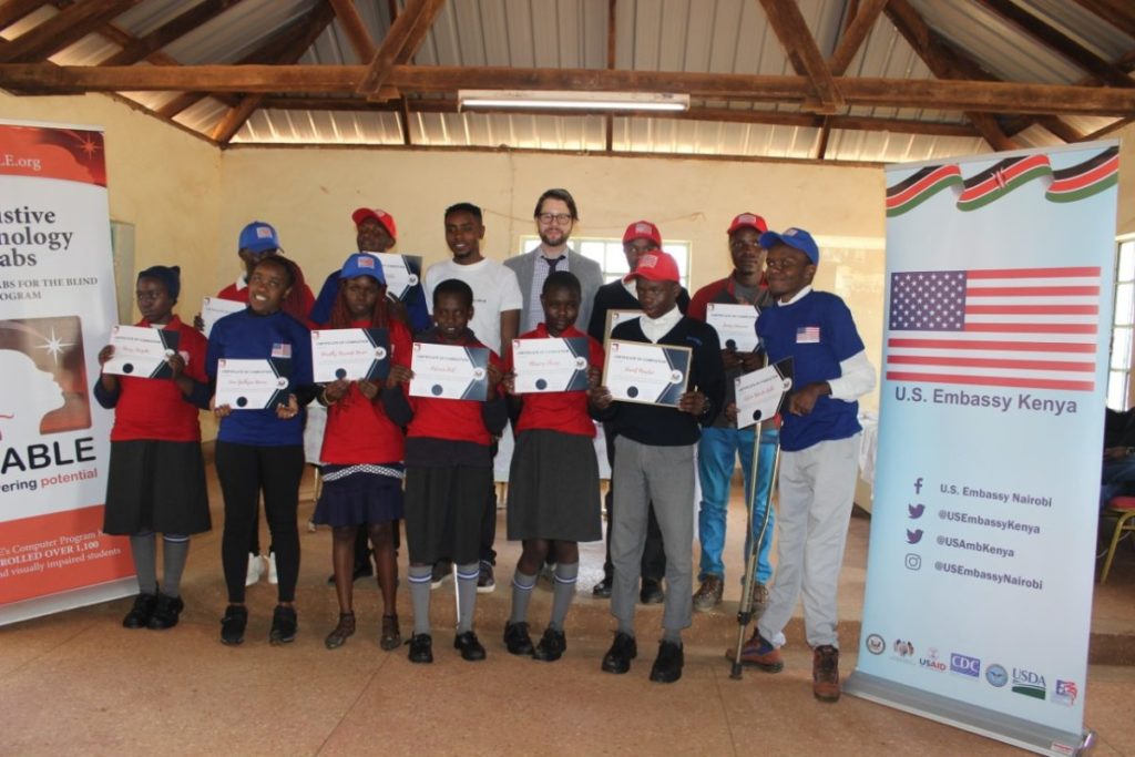 A group of Black students holding certificates look forward while they stand with a Black team leader in white shirt and a White man wearing glasses in a gray suit.