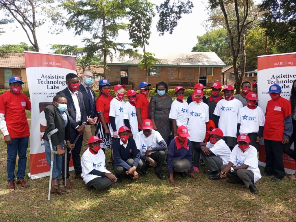 A group of students wearing red, white and blue USA hats, t-shirts and mask pose with guests and school administrator in front of their school and inABLE.org posters.