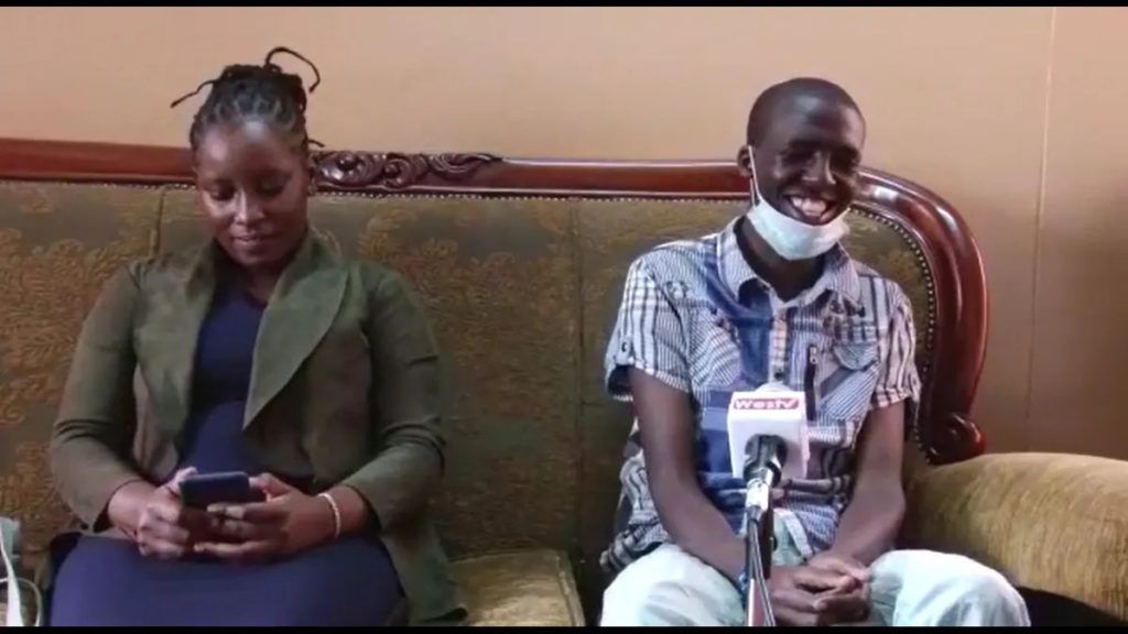 Eunice Kilungu on left wearing green jacket is seated on a couch looking at her iphone. Seated next to her on the right is a smiling Edwin Omulama with mask below his chin.