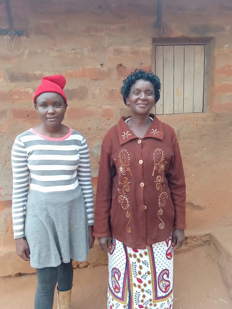 Facing forward on the left is Ruth Kasyoka, wearing a read hat and stripped dress. Next to her on the right is her mother Jacinta Nzeng’a, wearing brown jacket and colorful skirt. They are in front on red mud building.