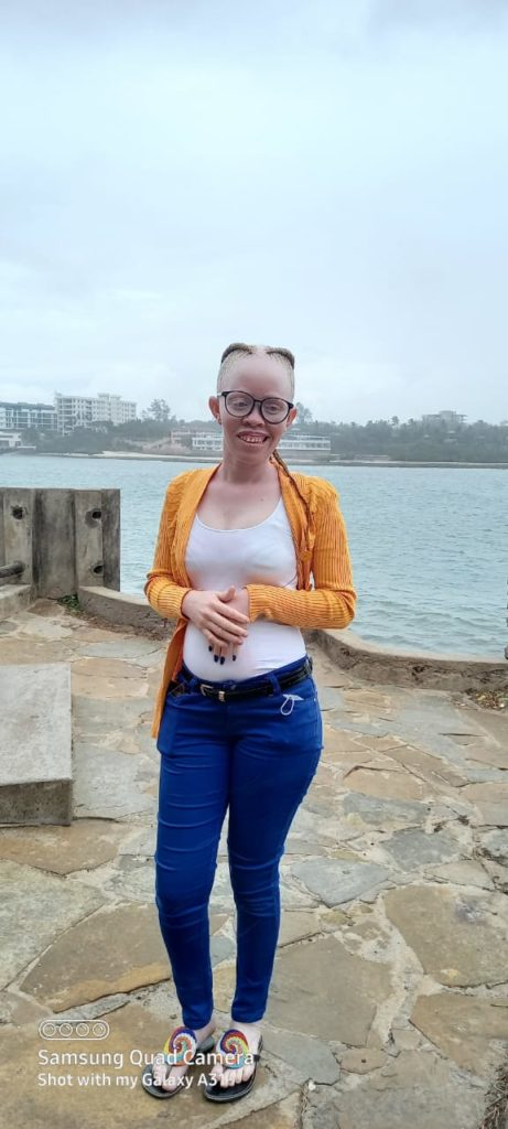 Fair skinned women, with braided hair, wearing glasses, an orange sweater, and white top stands near the ocean