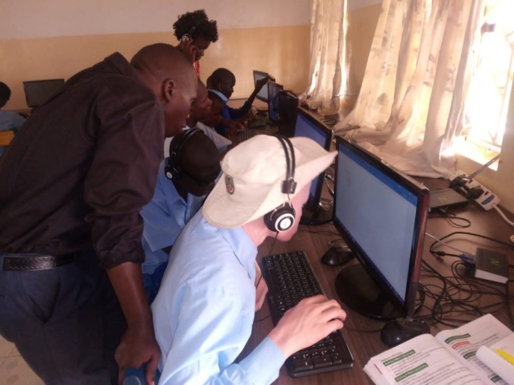 A black man wearing a back shirts looks at a computer screen over the shoulder of a student wearing a white hat, headphones and blue shirt .