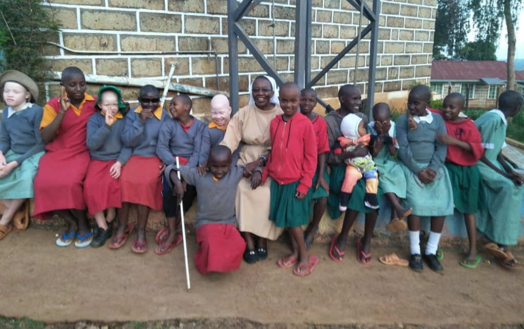 In the center of a group of joyful young students is a smiling Sr. Judith Felicity Khavwengesi. She wears a tan dress, white veil and glasses.