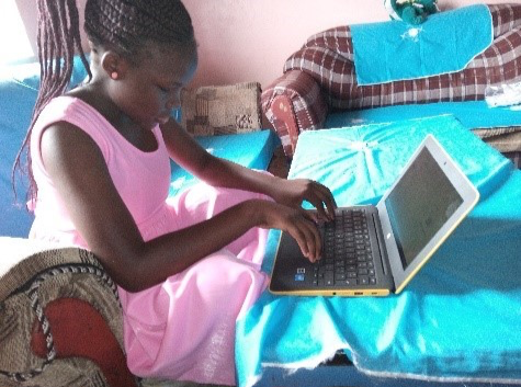 A blind girl wearing a pink dress and seated on a sofa is typing on her Chromebook laptop.