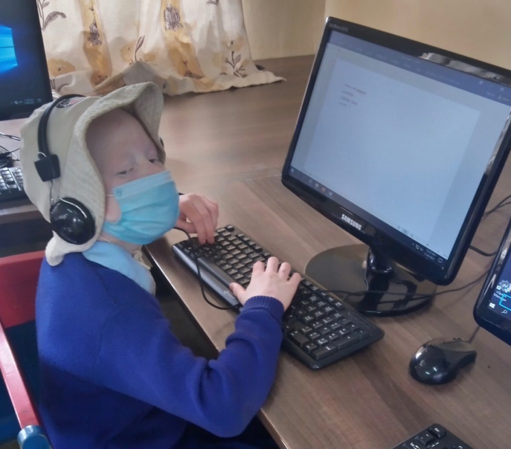 inABLE Student Joy Atieno types on a desk top computer. She wears a tan hat, headphones, blue sweater and looks to the right.