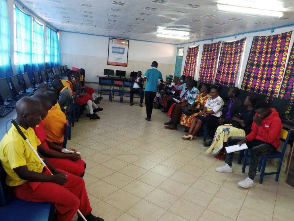 Black students wearing yellow and red school uniforms sit across from parents in computer lab