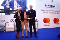 On a stage with blue carpet and an Inclusive Africa banner are a Brown women in tan braids wearing a black dress, next a Black man wearing glasses and holding a trophy, with a White man with white hair wearing a black suit standing on the right.