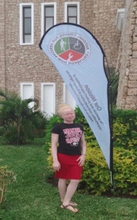 A women with light hair and skin wearing a black tee shirt and red skirt stands in front of a stone building with three windows above three white doors.