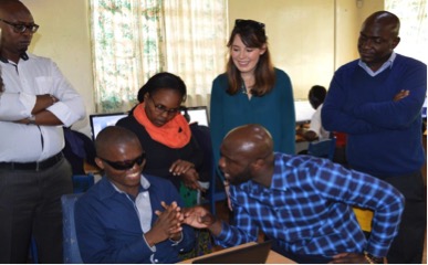 Mastercard representative Andrew Okuzuwa , who is seated to the right of inABLE intern Anthony Wambuwa talks while Anthony smiles and four onlookers - Joe Kiarie, Brenda Kiema, Kaylee Stewart and George Siso- stand behind them watching the interaction.