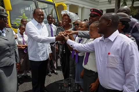 Kenya President in front of yellow school bus given to Likoni High School for the Visually Impaired in Kenya joins hands with a group of onlookers standing in front of him.