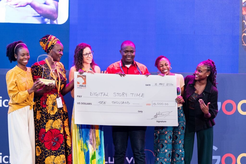 A picture of the Digital Story Time Team including Georgine Auma and Laura Allen from Google. They're all on stage holding onto a dummy cheque written Digital Story Time, Ten Thousand Dollars. They're also holding a trophy and smiling.