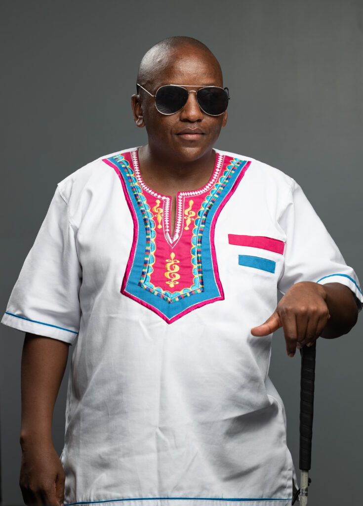 A picture of Mr. Tap standing and posing for a picture. He's wearing black sunglasses, a white shirt with colored prints near the collar. He's using his right hand to hold onto his white cane.