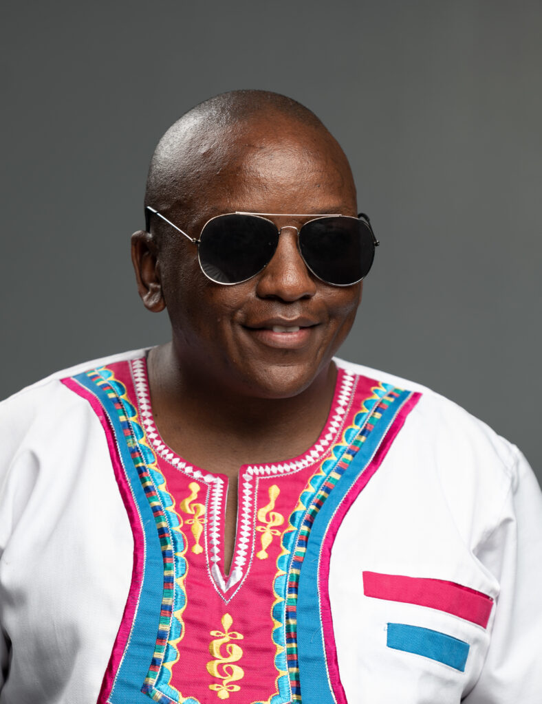 A passport picture of Mr. Tap standing, smiling and posing for a picture. He's wearing black sunglasses, a white shirt with colored prints near the collar. 