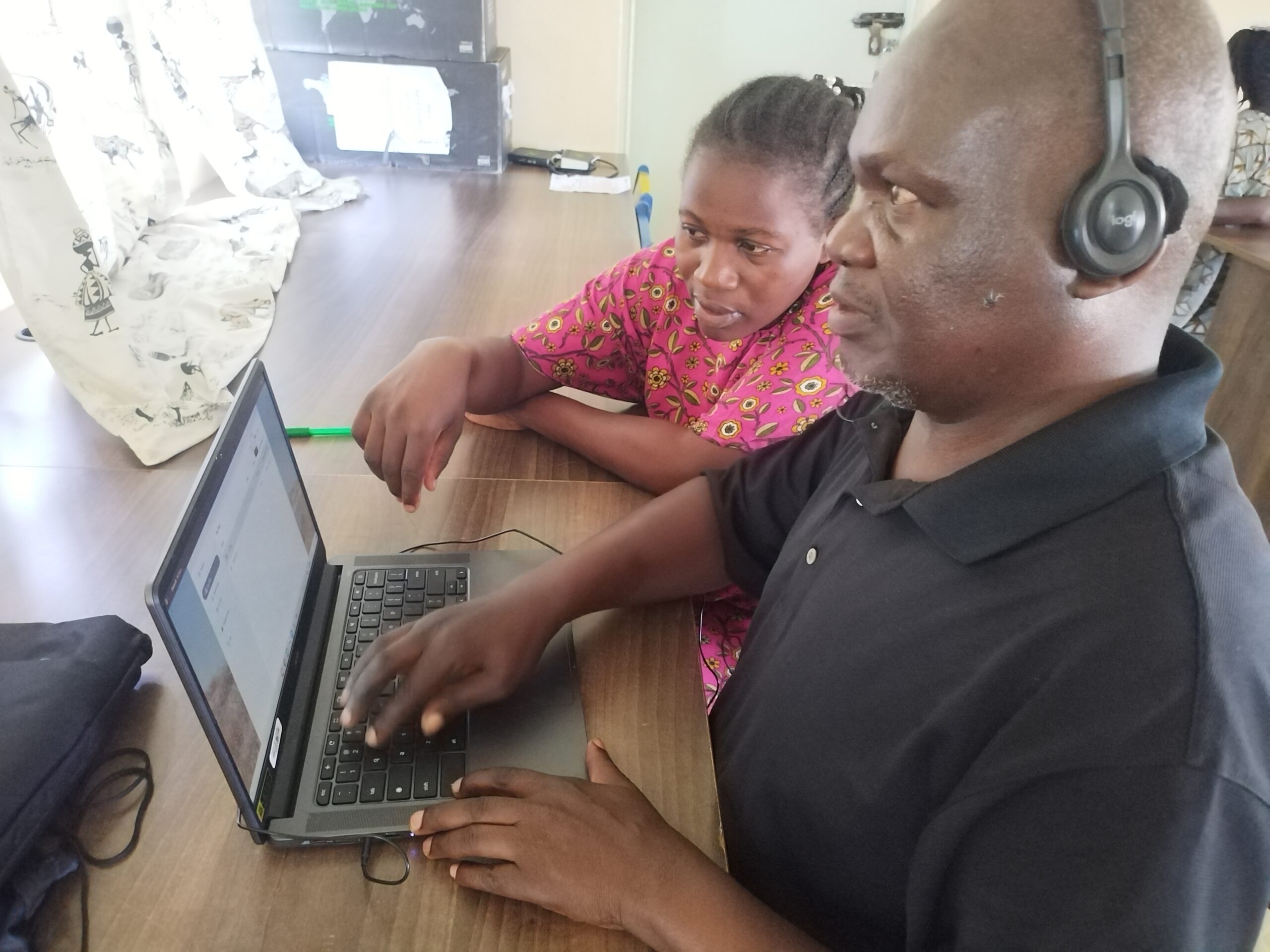 A picture of Mr. Fredrick Ochieng using a laptop and wearing headphones. There's a lady seated next to him.