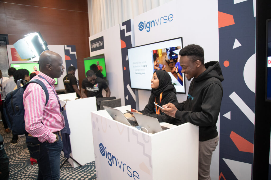 A picture of Sewu-Steve Tawia talking with Elly Savatia and Gheida. Elly and Gheida are standing behind a Signvrse branded booth inside the AT Innovation Village 2024. There are other innovators and attendees standing inside the AT Innovation Village.