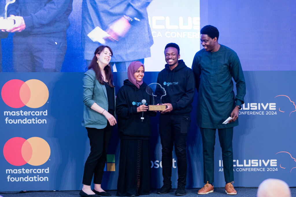 A picture of Elly Savatia and Gheida smiling, standing on the Inclusive Africa conference stage and holding the 1st Runners Up Digital Accessibility and Assistive Technology Innovations Award. Two representatives from the Mastercard Foundation are standing next to them.