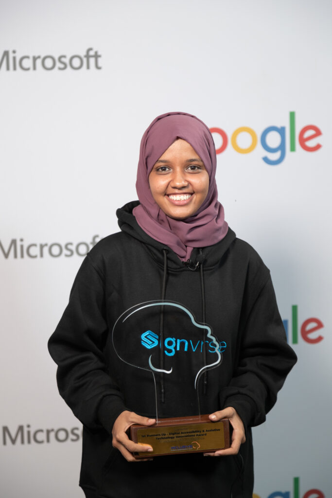 A picture of Gheida smiling and standing infront of a branded booth. She's wearing a branded Signvrse black hoodie. She's holding an award written Signvrse, 1st Runners Up Digital Accessibility and Assistive Technology Innovations Award. 5th Inclusive Africa Conference Conference