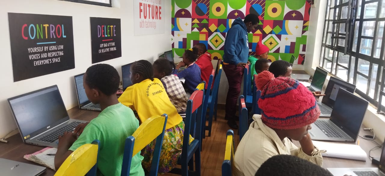 A picture of students using Chromebook laptops during a coding class. The students are seated down while a teacher is standing up.