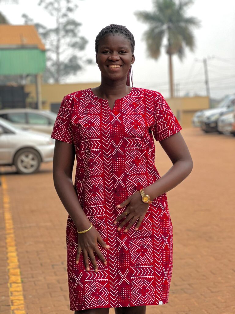 ​​This is a picture of Gloria smiling and posing for a photo. She has black braided hair and is wearing a red and white patterned dress, a gold bracelet, and a gold watch.