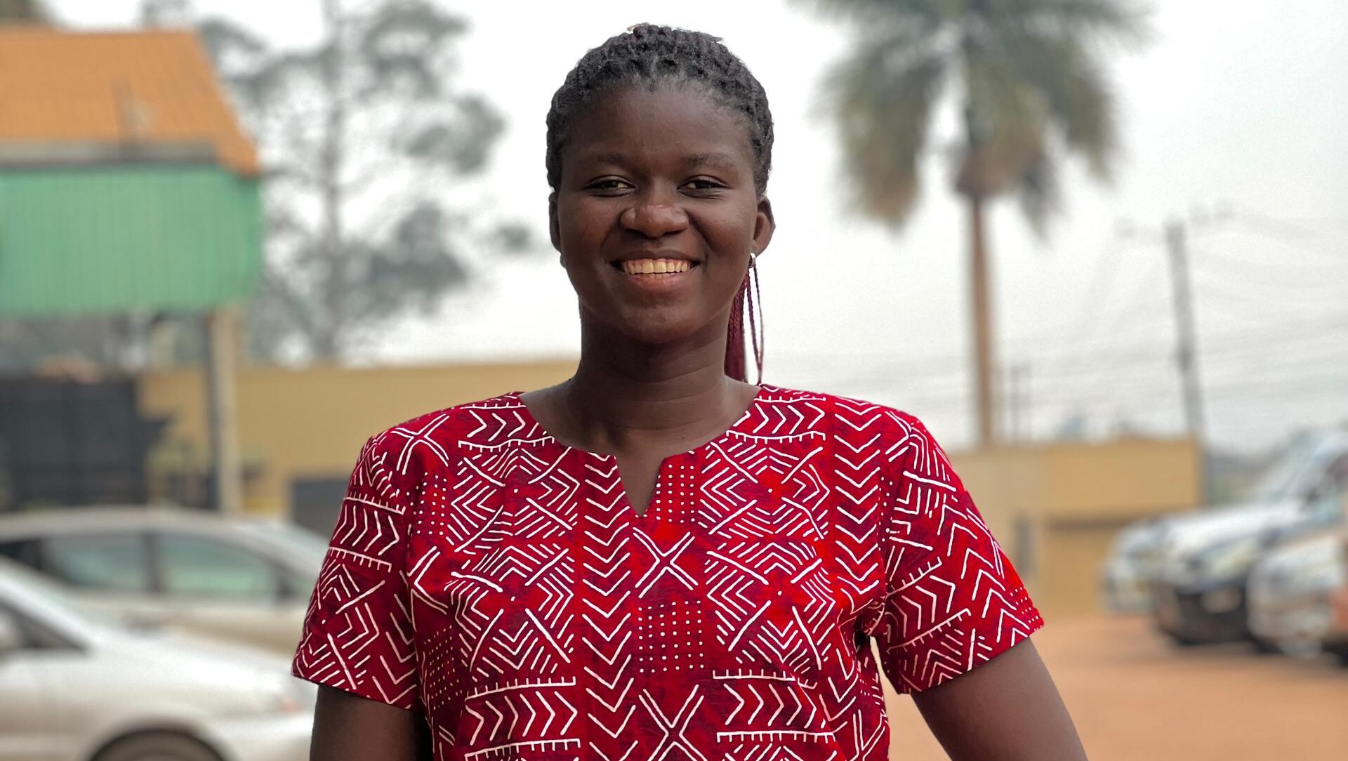 ​​This is a picture of Gloria smiling and posing for a photo. She has black braided hair and is wearing a red and white patterned dress, a gold bracelet, and a gold watch.