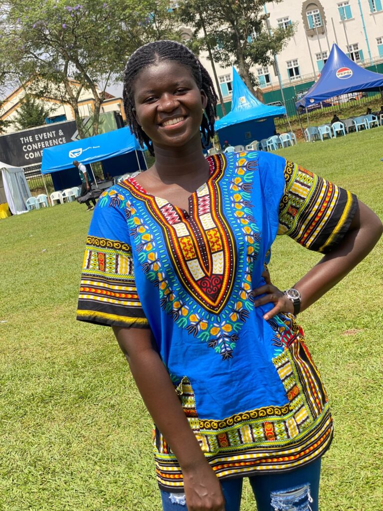 ​​This is a picture of Gloria smiling and posing for a photo. She has black braided hair and is wearing a blue multi-coloured dashiki and blue jeans.