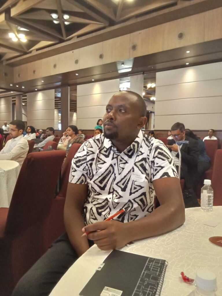 A picture of Anthony Njoroge seated on a chair, holding a pencil and listening keenly. He's wearing a shirt with black and white patterns plus black trousers. There are other people seated at different tables in a large room during the Zero Project Technology Forum