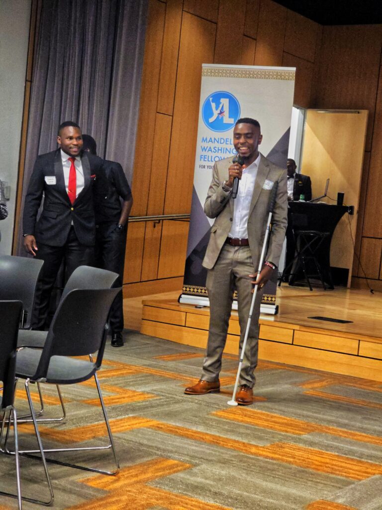 A picture of Julius Mbura standing infront of a Mandela Washington Banner, holding a microphone in his right hand and smiling. He's wearing a light blue shirt, brown suits and dark brown shoes. He's holding his white cane with his left hand.