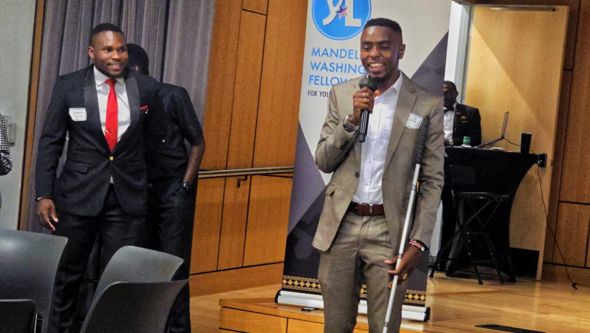 A picture of Julius Mbura standing infront of a Mandela Washington Banner, holding a microphone in his right hand and smiling. He's wearing a light blue shirt, brown suits and dark brown shoes. He's holding his white cane with his left hand.