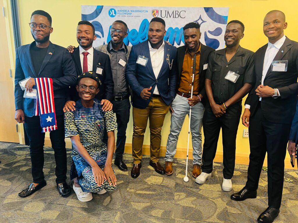 A group picture of men, including Julius Mbura, smiling and posing for a photo. Julius is wearing light grey pants, a dark brown shirt and a black coat and holding his white cane with his left hand 