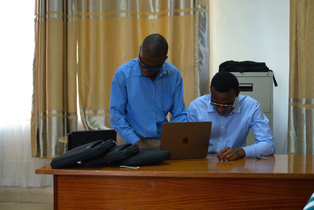 A picture of Nahashon Kimochu seated down with a Macbook on a table. Wambua, an inABLE instructor is standing near him.