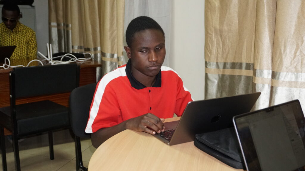 A picture of Marvin Masiali seated down and using a Macbook laptop on a table. He's wearing a red shirt with a white stripes and a black collar.