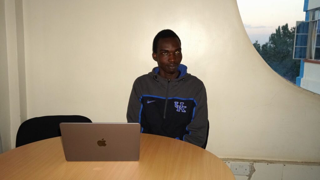 A picture of Philip Omele seated down on a chair with a Macbook laptop on a table near him. He's a grey and black hoodie.