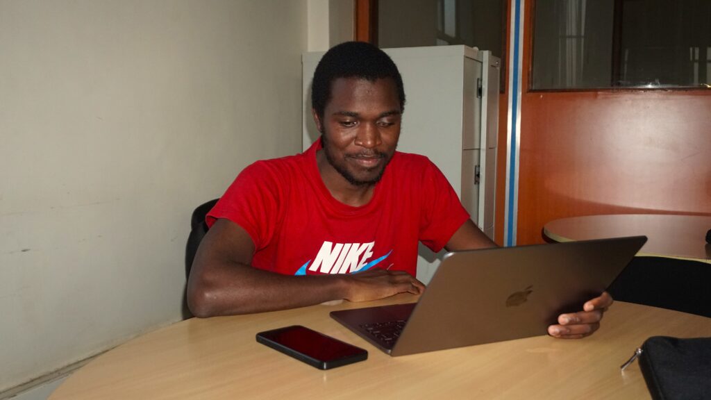 A picture of Elvis Apondi Odury seated down and using a Macbook laptop. He's smiling and wearing a red t-shirt.