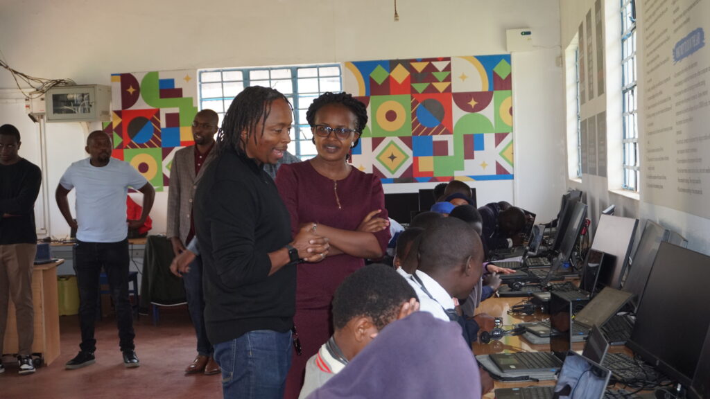 A group of adults are standing observing students seated and working on their Chromebooks, demonstrating the coding skills they have been learning.