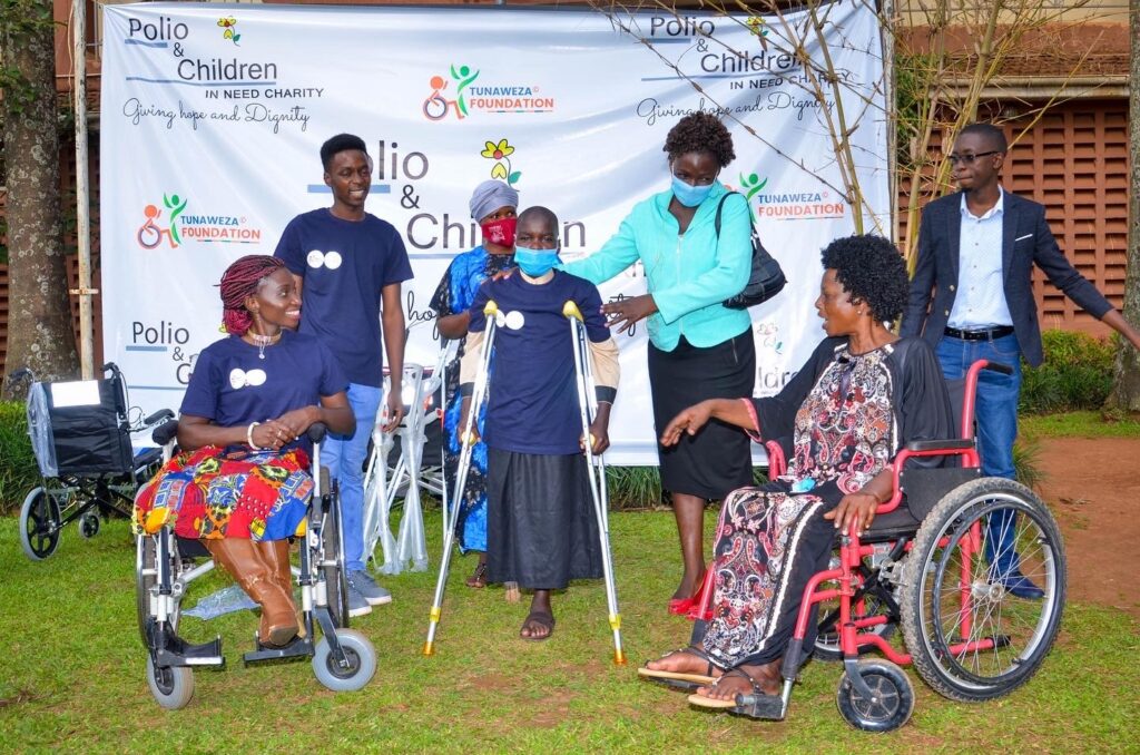 A group photo showing individuals with disabilities including Victo Nalule and their supporters during a community event. Two women in wheelchairs are smiling, while a young boy with crutches stands in the middle, supported by a woman in a green blazer. The backdrop features logos for "Polio & Children in Need Charity" and "Tunaweza Foundation," with the tagline "Giving Hope and Dignity."