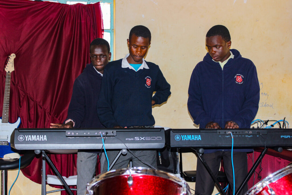Three male students in school uniforms play on Yamaha keyboards in a classroom setting. They appear focused on their music, with a red curtain and a guitar in the background. A drum set is partially visible in the foreground. The students are wearing navy blue sweaters with a red crest emblem, indicating they belong to the same school or institution. The room has plain cream-colored walls and a window letting in natural light.