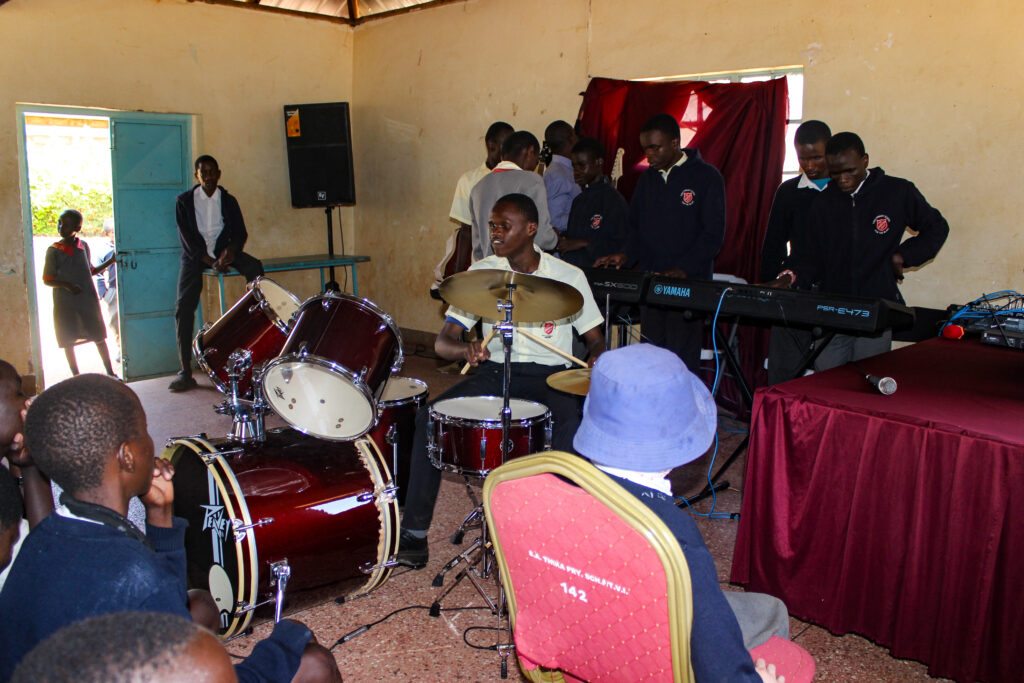 A group of students in school uniforms are performing music in a classroom. One student is playing a red drum set, while others are playing Yamaha keyboards and guitars. Several students are seated and watching the performance. The classroom has a simple setup with a wooden ceiling, cream-colored walls, and a red curtain behind the performers. A door is open, letting in natural light, and a few students are standing near the entrance.