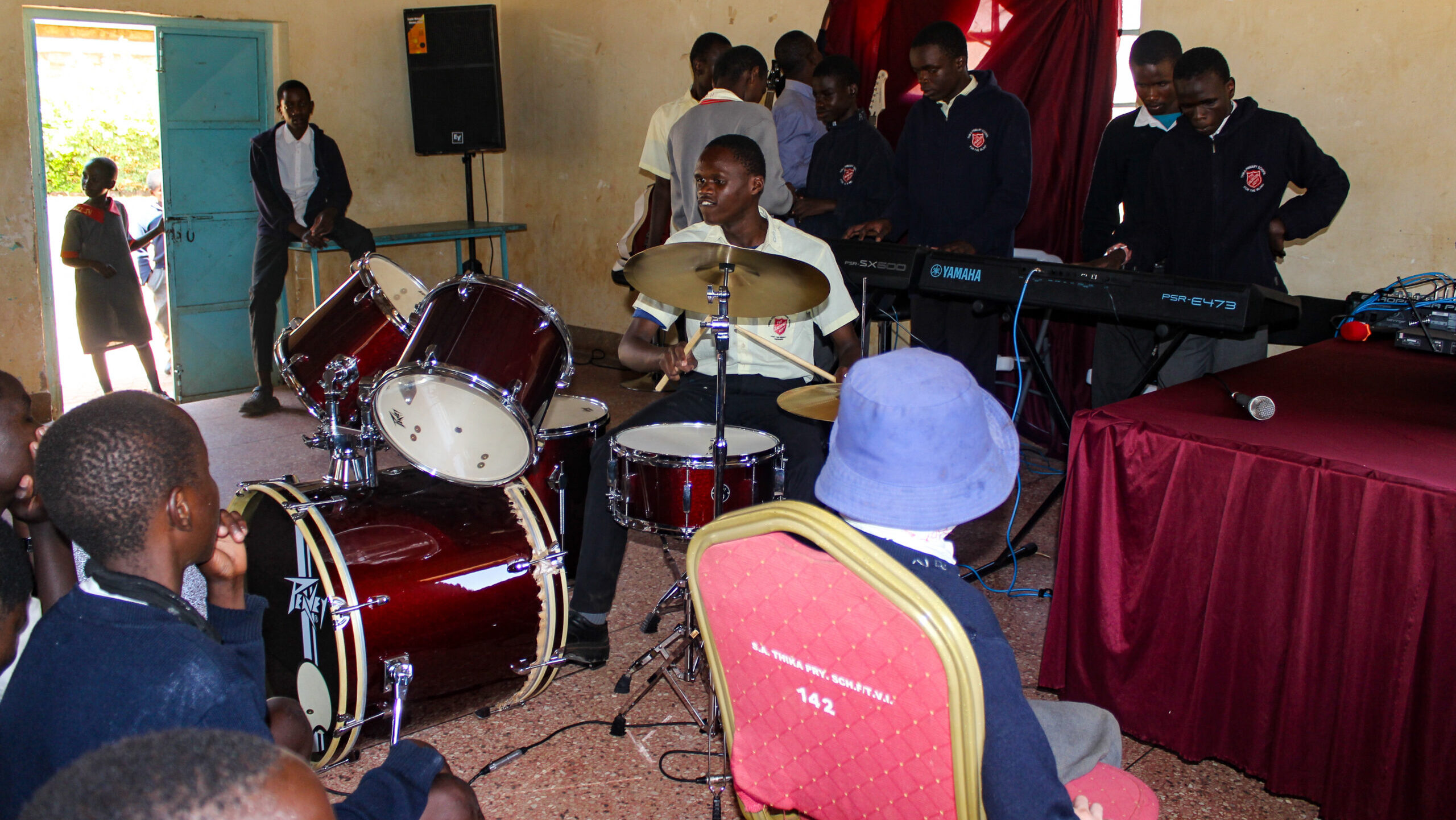 A group of students in school uniforms are performing music in a classroom. One student is playing a red drum set, while others are playing Yamaha keyboards and guitars. Several students are seated and watching the performance. The classroom has a simple setup with a wooden ceiling, cream-colored walls, and a red curtain behind the performers. A door is open, letting in natural light, and a few students are standing near the entrance.