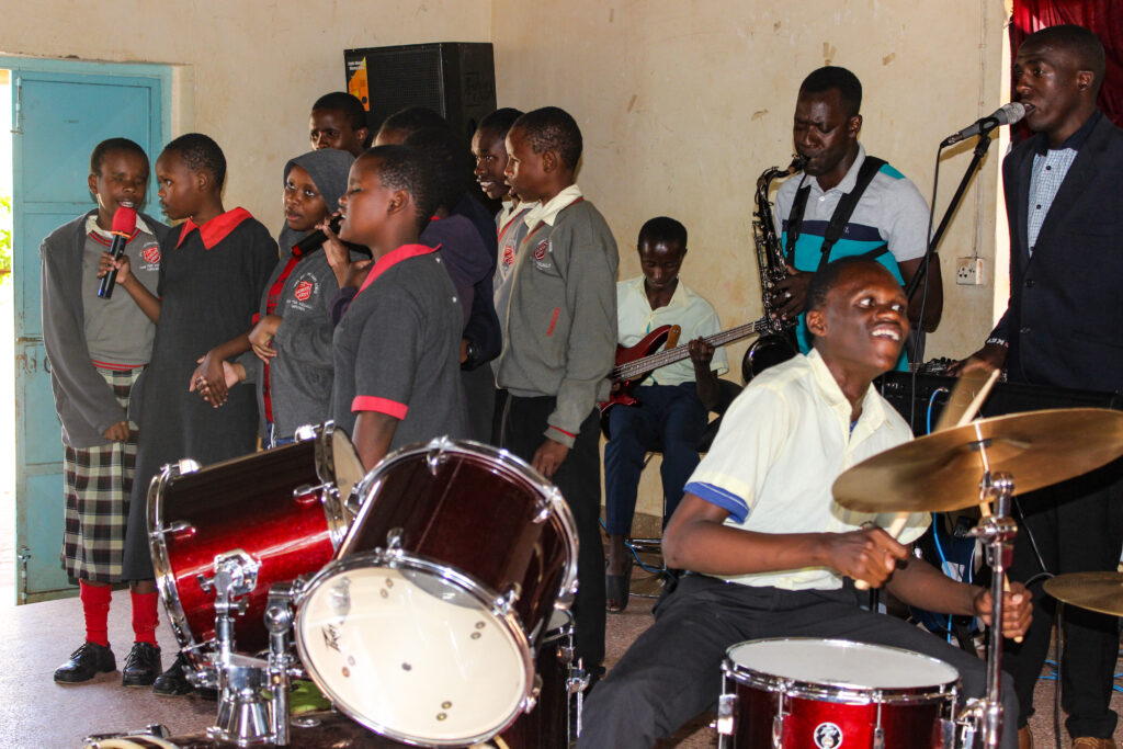 A group of students in school uniforms are performing music in a classroom. One student is playing a red drum set, while others are playing Yamaha keyboards, saxophone and guitars. There are female students who are standing near each other and singing.