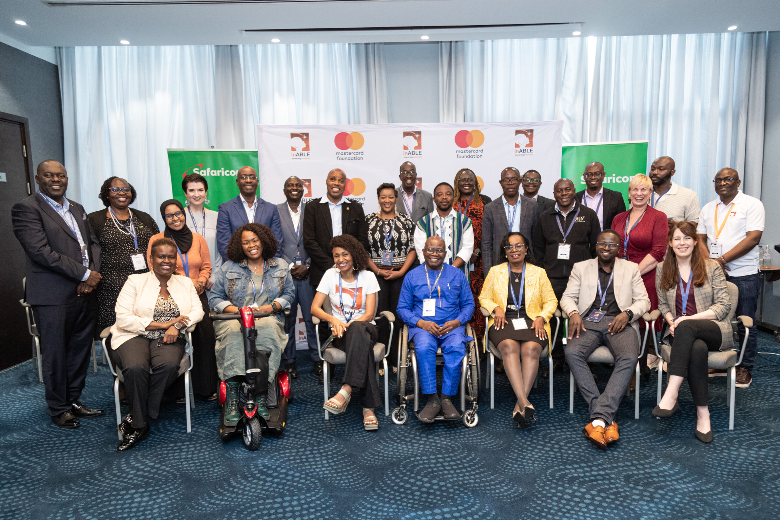 A group picture that includes Catherine Wamwangi, Lizzie Kiama, Irene Mbari-Kirika, Kojo Tetteh, Tabie Kioko, Kylee Stewart, Sarah Ford, Justice Laibuta, Joe Kiarie, George Siso and other people smiling and posing for a picture. Behind them, there are banners with logos for Safaricom, Mastercard Foundation and inABLE.